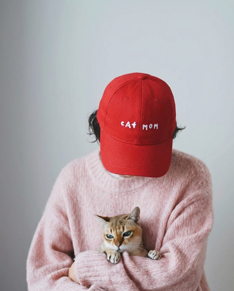 A cat mom in a red hoomans CAT MOM cap and a pink sweater, holding her tabby cat