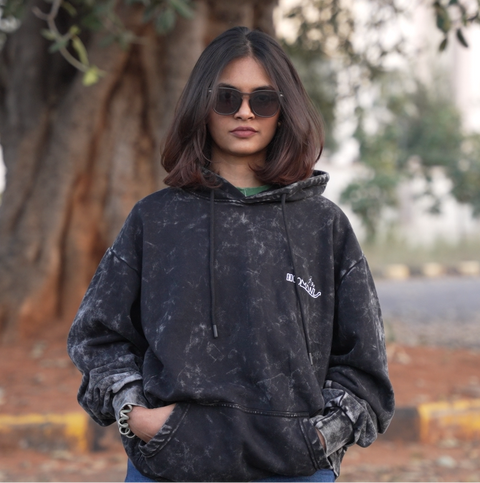 A person wearing a black acid wash hoodie with a Hoomans brand logo, standing outdoors with trees in the background.