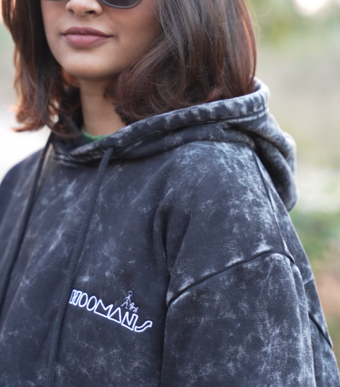 Person wearing a black acid wash Hoomans hoodie with a hoomans brand logo, outdoors.