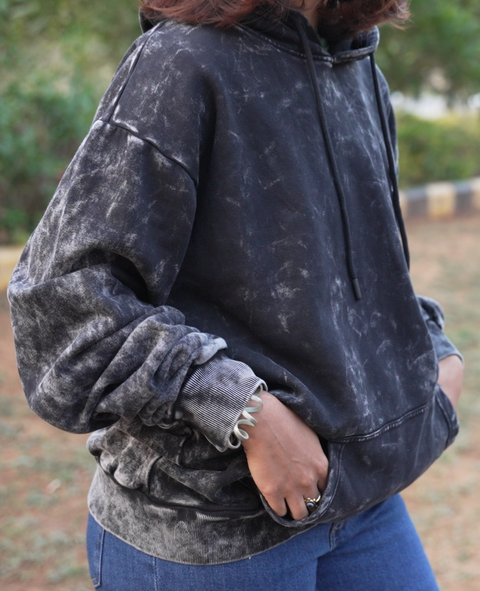 Person wearing a black acid wash hoodie outdoors with greenery in the background