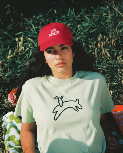 Person wearing a red cap with 'Dog Person' text and a light green t-shirt with a dog illustration, sitting in grass.