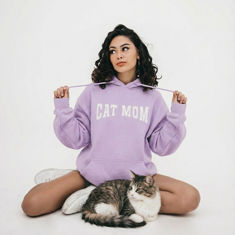 Woman wearing a purple 'CAT MOM' hoodie sitting with a cat on a white background