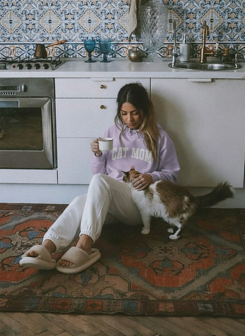 Woman sitting on a rug in a kitchen with a cat, holding a mug, wearing Cat Mom Hoodie.