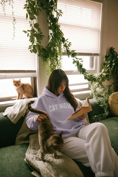Woman reading a book with two cats on a couch in a cozy room, wearing Cat Mom Hoodie.