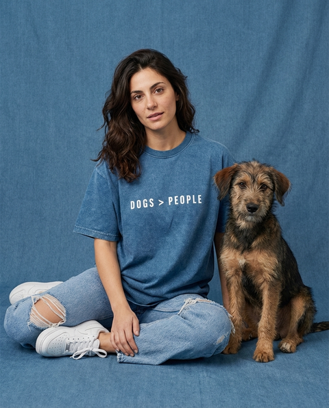 Woman wearing a denim blue oversized t-shirt with 'DOGS > PEOPLE' text, sitting next to a dog on a blue background