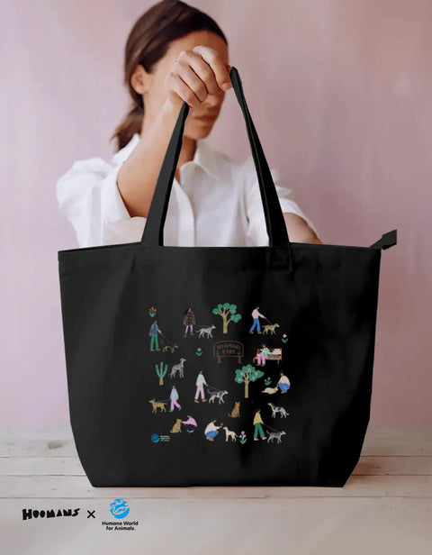 Dog Park Night Scenes black zip tote bag held up, printed with an illustrated dog park of dogs, people and a HOOMANS PARK board, Hoomans x Humane World for Animals India lockup