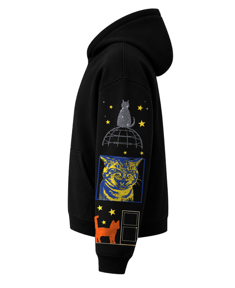 Left side of the black boxy hoodie: a sparkling cat on a globe, a blue and yellow hissing tabby in a frame and an orange cat at a door, with yellow stars down the sleeve
