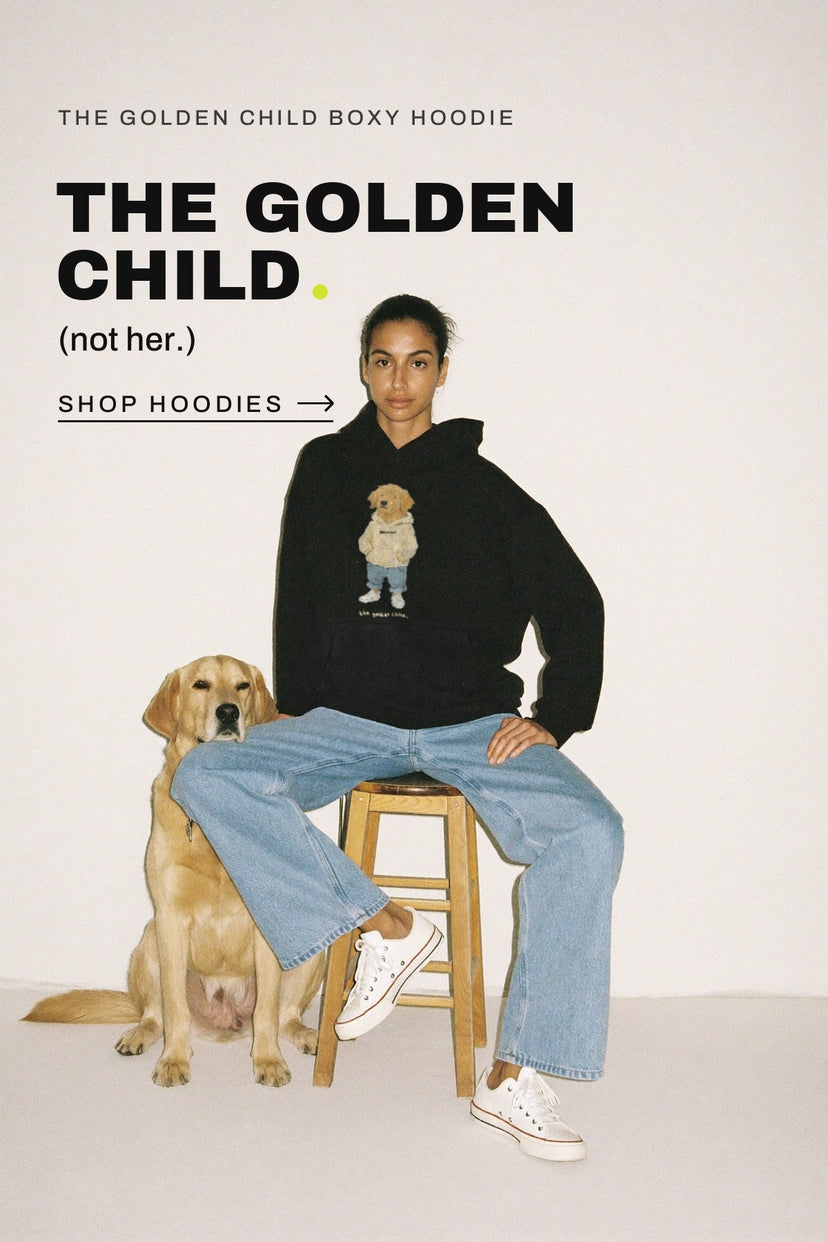 A woman in the black Golden Child boxy hoodie sits on a wooden stool beside her golden retriever. The golden child. (not her.) Shop hoodies.