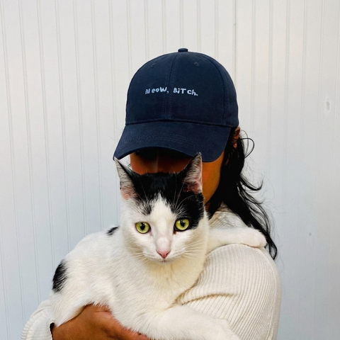 Person wearing a navy blue cap with 'Meow, Bitch' text, holding a black and white cat against a light background.